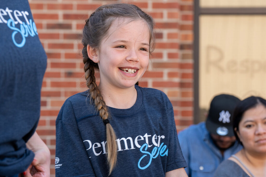 A child smiles while wearing a "Perimeter Serves" t-shirt